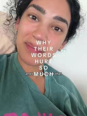 A selfie-style shot featuring a woman with dark, curly hair. She's facing the camera, smiling slightly, eyes looking upwards. Text overlays in a vertical stack that reads "WHY THEIR WORDS HURT SO" and "and I might wait for that". She is wearing a teal t-shirt. The background has a cream colored bed and pillow.