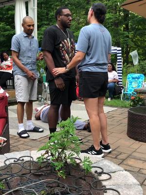 The image captures a medium outdoor scene during the day, likely a backyard gathering. The focal point is a group of people involved in a moment, with one individual's hands placed on another's head. A third person stands to the left, observing the scene. In the background, other people are present, suggesting an event or celebration. The overall impression is that of a casual social gathering. The color palette is natural, with greens from the surrounding foliage and variations of light and dark tones on the people’s clothes and skins.