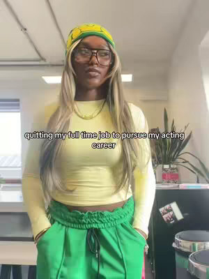A woman is centered in a medium shot, standing in what seems to be an office or studio setting. She wears a yellow, long-sleeved crop top paired with green track pants featuring a drawstring. Her outfit is complemented by a yellow and green patterned beanie, large-framed glasses, and long blonde-highlighted hair. She has her hands in her pockets, and the image has natural lighting. Behind her, a plain white wall and visible ceiling lights suggest an indoor environment. Text overlayed at the bottom reads, \"quitting my full time job to pursue my acting career\".