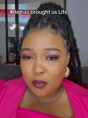 A close-up shot of a woman with dark, curly hair pulled back in a high ponytail. She wears heavy eye makeup, including pink and berry shades. Her lips are a deep berry color. The background is blurred, suggesting an indoor setting. A text overlay reading 'He has brought us Life' is over the image.