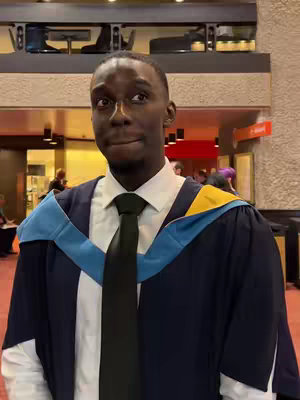 A young Black man in graduation attire smiles, looking off to the side. He wears a dark tie, a white shirt, and a graduation gown. The background shows a hallway with signs and other people, suggesting a university setting.