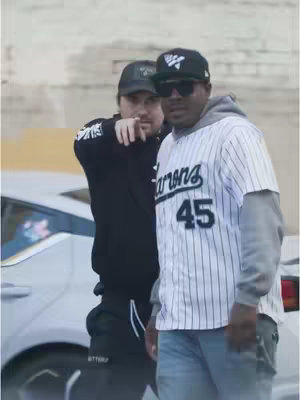 Two men are standing outside. The man in the front is wearing sunglasses, a black hat, and a white baseball jersey with the number 45 on it. The man behind him is wearing a black hat and a black jacket. A silver car is partially visible in the background.
