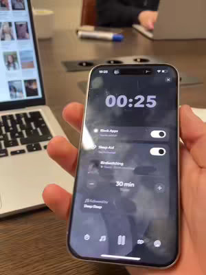 A hand holding a smartphone displaying an app interface. The phone is centered, and the background shows a laptop and parts of a desk, the phone's screen shows a timer and app controls.