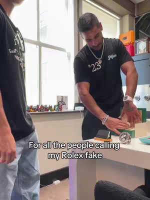 Two men are in a room, one showing a Rolex watch on his wrist while the other stands nearby. The scene is brightly lit suggesting a professional environment with some decorative items in the background on a counter. Text overlaid says, "For all the people calling my Rolex fake" in white.