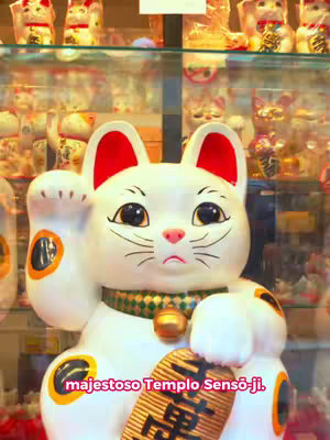 The image shows a close-up of a large, white ceramic Maneki-neko cat. It has a smiling face, big black eyes, red ears, and a green patterned collar with a gold bell. The cat is holding up its left paw. Behind the cat, there are shelves filled with other Maneki-neko cats and various merchandise. Text overlayed on the image reads: "majestoso Templo Sensō-ji."
