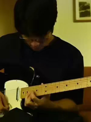 A person wearing a black top is playing an electric guitar. Their face is partially obscured by their head and a pair of glasses. The guitar's body is white with a black pickguard, and the neck is a light-colored wood.
