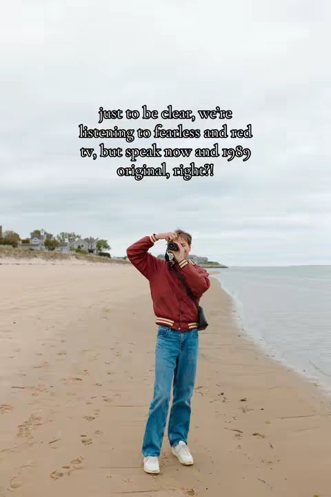 A person stands on a sandy beach taking a photo. The person is dressed in a red jacket, blue jeans, and white sneakers. Behind the person is the ocean and sky.