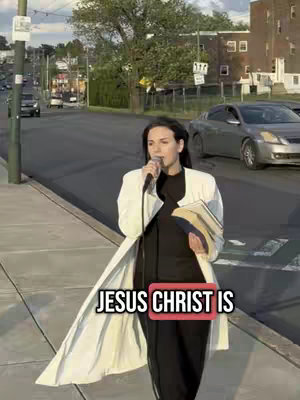 A woman speaking into a microphone on a sidewalk, holding a book. The background shows a street with cars and buildings.