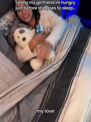The cover image shows a person lying in a bed, partially covered by a blanket. The person is holding a white teddy bear and a phone in their arms, and is smiling. The text overlayed at the top says 'Telling my girlfriend im hungry just before she goes to sleep..' The text at the bottom says 'my love'.