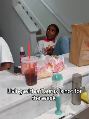 A young Black woman sits indoors, looking upwards with a slight smile, holding a sandwich. Her surroundings include a counter with food items, a stairway in the background, and a clear drink with a straw. The composition gives a casual, domestic feel.