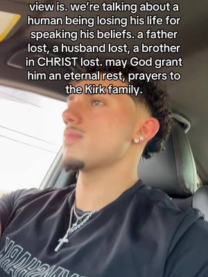 A young man with short dark hair and a goatee is filmed from the chest up, partially inside a car. He is wearing a dark T-shirt with a cross necklace. Text is overlaid.