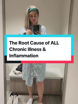 A woman is seen in a mirror, taking a selfie. She has blonde hair, a green headband, and is wearing a light blue button-down shirt or dress. A text overlay in a rectangular shape declares "The Root Cause of ALL Chronic Illness & Inflammation." The background shows a minimal decor with a white cabinet with two pairs of shoes, with a simple tan wood color, emphasizing the focus on the speaker.