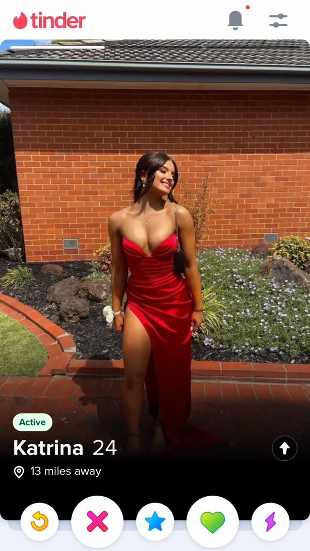 A woman is featured on a dating app profile, standing in front of a brick wall and some shrubbery. She is wearing a low-cut red dress with a high slit. On the left side, there's a patch of green grass with some rounded boulders. The app interface shows her name, age, and distance.