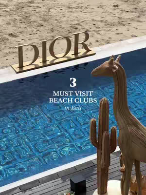 A cover image showing a swimming pool with a high-end brand's sign floating above. The pool reflects the clear blue sky, and in the foreground, a giraffe statue and a cactus enhance the high-end vacation vibe with text overlayed which says "3 MUST VISIT BEACH CLUBS in Bali"