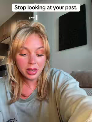 A woman with blonde hair and light makeup is looking directly at the camera, with a focused expression. She has rosy cheeks and is wearing a light green t-shirt and a gold necklace. The background includes glimpses of a kitchen setup to the left and a dark decorative square artwork on the right.