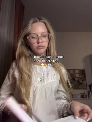 A young woman with long blonde hair and glasses is holding a book and looking at the camera. Behind her is a room with a curtain in the background.