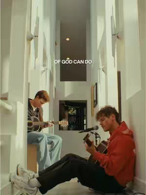 Two young men playing acoustic guitars in a well-lit hallway. One is seated, playing a guitar into a microphone, while the other sits on a bench, also playing. The image is vertically oriented, focusing on the musicians and the space around them.
