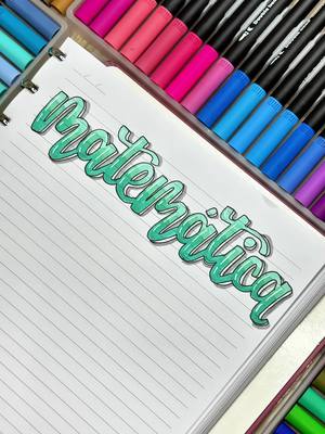 The image showcases a notebook with the word "Matemática" (Mathematics) written in a vibrant, three-dimensional style using turquoise brush pens. The title stands out against the lined paper of the notebook. In the background, a collection of colorful brush pens is arranged, adding a visually appealing layer.