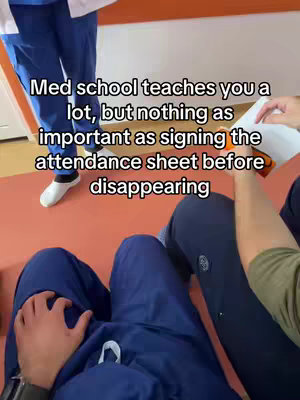 The image shows a scene from a medical school setting. The primary figures are two individuals, possibly medical students or professionals, wearing blue scrubs. The composition is a medium shot with a point-of-view perspective. The background includes a segment of an orange wall against a brown platform. The text overlay reads: Med school teaches you a lot, but nothing as important as signing the attendance sheet before disappearing