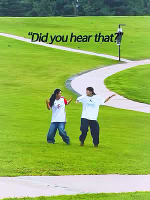 Two people are on a green grassy field under an overcast sky. A winding path leads away from the foreground. One person is wearing a white t-shirt, black pants, and a hat. The other person is wearing a white shirt, blue jeans and a shirt with red and white elements. Text "Did you hear that?" is in white at the top of the frame.