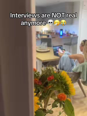 The image shows a woman sitting in a room with a laptop. The text overlay reads "Interviews are NOT real anymore". A bouquet of flowers is in the foreground, obscuring part of the view. The background shows parts of the room, including a kitchen area. The composition suggests a casual, indoor scene focusing on work or content creation.