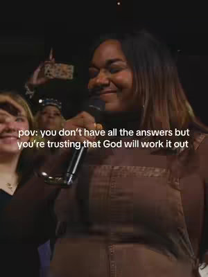 A medium shot captures a Black woman smiling with a microphone in hand. She's wearing a brown jumpsuit and facing slightly right, with her face and upper body in focus. Behind her are other individuals, partially visible. Text overlayed on the image reads: "pov: you don't have all the answers but you're trusting that God will work it out". The lighting is slightly dim, suggesting an indoor setting.