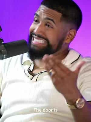 A close-up shot of a man with a beard and short dark hair. He's wearing a white shirt with black trim, and a gold watch. He's gesturing with his right hand and appears to be speaking with enthusiasm. The background is a bright, blurred purple.