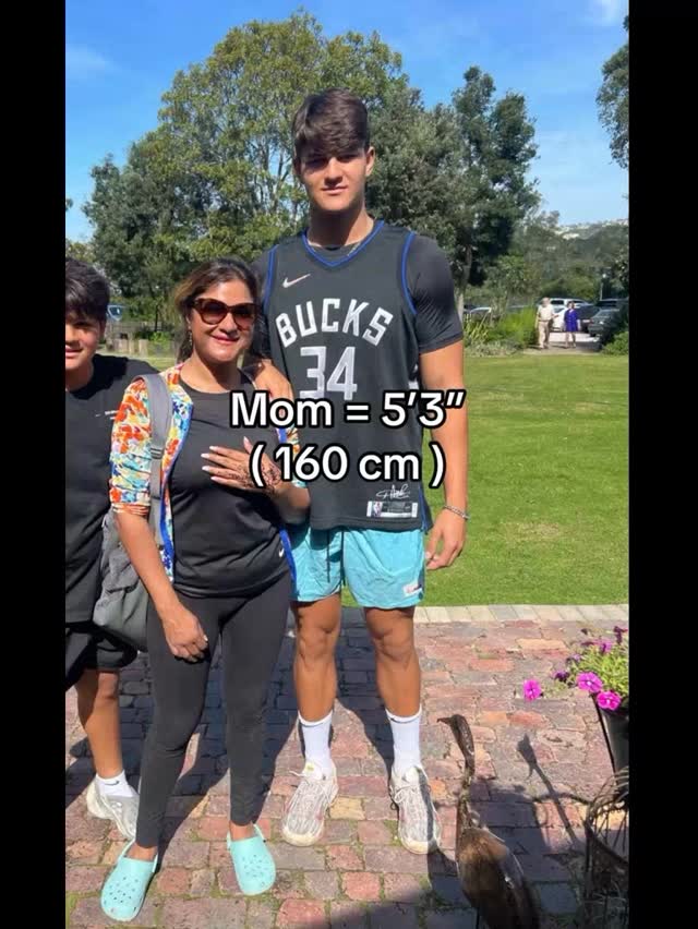 A family of three, mother, son, and another son stand outside in front of a tree. The taller son wearing a basketball jersey.