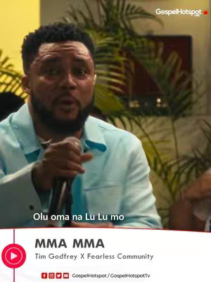The cover image features a man speaking into a microphone in a well-lit setting. The man has short, dark hair and a trimmed beard. He is wearing a light blue blazer, and the background includes blurred green foliage and a dark-red object. The video title area showcases the text "MMA MMA," alongside the artist's name and the "GospelHotspot" logo.