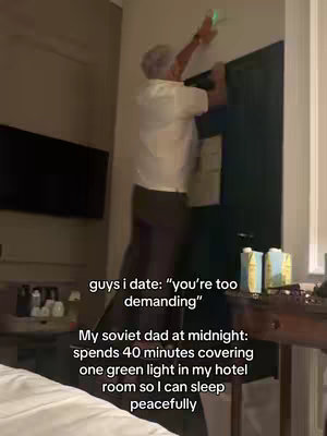 A casually framed shot with text overlay. The image features a man in a hotel room, clad in a white shirt and dark pants, standing on a small step, his back to the camera as he reaches upward, presumably to adjust something on the wall. The background is dimly lit, revealing parts of the room's decor, including a television and a table with items.