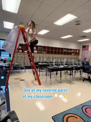 A woman is standing on a red ladder in a classroom, reaching towards the ceiling, where a plastic film is being installed. The classroom has desks, chairs, and cabinets along the back wall, with an American flag hanging on the right corner