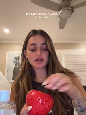 The cover image features a woman looking directly at the camera, seemingly talking. She is holding a large, red, strawberry-shaped decorative object. The background shows a simple room with a ceiling fan, and a framed image is partially visible behind her.
