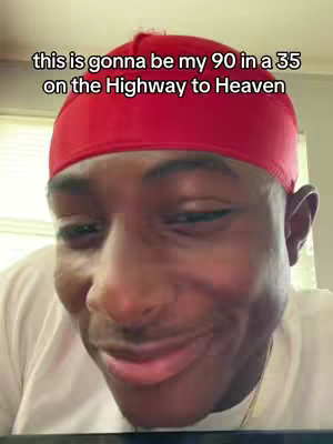 A close-up shot features a Black man, positioned from the chest up, with his face taking up most of the frame. He wears a bright red durag and a white t-shirt. His expression is animated with a slight smile, and a sense of playful joy. The background is out of focus, with a plain wall visible, putting all the focus on the subjects face. The image is well-lit, highlighting his facial features.