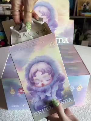The image shows a person's hand opening a POP MART box that contains a Skullpanda L’impressionnisme blind box. The box and packaging are primarily pastel purple and pink, featuring an illustration of the Skullpanda character with closed eyes, wearing a fuzzy hooded cloak. The person opening the box has long nails painted with a green design. The background shows several boxes stacked, and text on the opened box reads “SKULLPANDA” and “L’impressionnisme”.