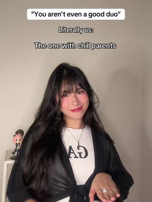 A woman with long dark hair and bangs stands in front of a plain background. She is wearing a white t-shirt with a black design and a partially buttoned black jacket. Text overlays are present at the top, indicating a caption.
