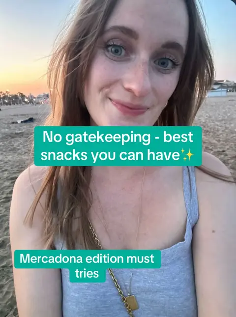 A selfie taken on a beach at sunset. The creator, a woman with long, wavy hair, smiles towards the camera. Text overlays in a turquoise box promote the video theme: "No gatekeeping - best snacks you can have" and "Mercadona edition must tries". The background shows the beach, the sea, and buildings in the distance.