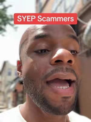 A close-up shot of a Black man with a shaved head and a beard, speaking with an open mouth. A red rectangular text overlay at the top reads "SYEP Scammers". The background is blurred but appears to be an outdoor setting.