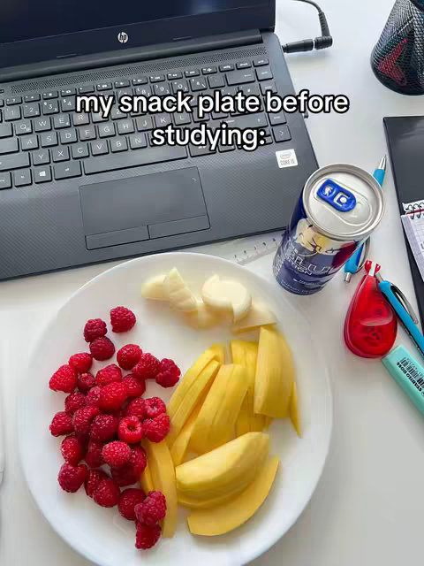 The image shows a plate of snacks next to a laptop. On the plate are raspberries, sliced mango, and other fruits. A can of what appears to be an energy drink and a few other items, such as pens and a sharpener round out the image.