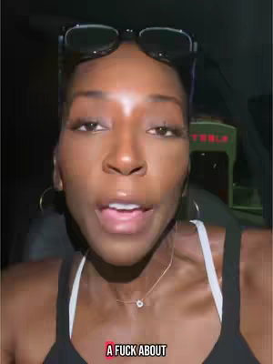A selfie-style shot of a woman inside a vehicle. She is looking directly at the camera with an open mouth, wearing a black top and a necklace. Her hair is styled back, and glasses are perched on her head. An in-car dashboard and a Tesla charging station are visible in the background.
