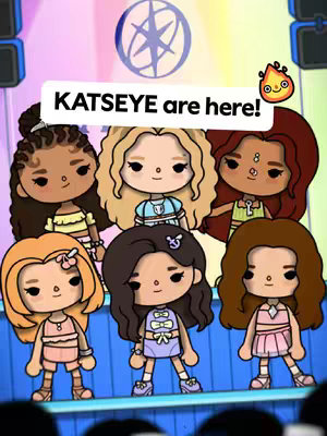 The cover image features six stylized cartoon figures of girls, arranged on a stage-like setting. Above, a banner reads 'KATSEYE are here!' in a white font against a black shape. A fire-like cartoon character is to the right of the banner. The cartoon girls vary in skin tone and hairstyle, each with unique clothing, standing and posing on a stage with blue and green lights.
