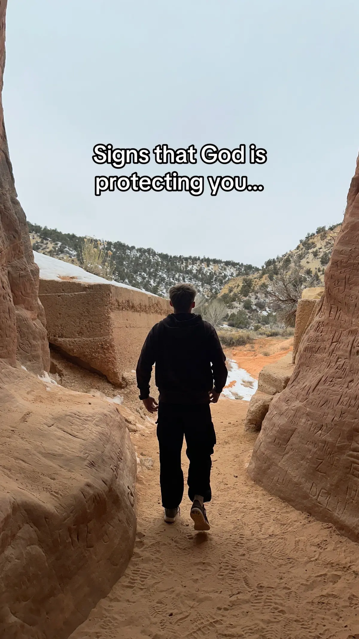 A person walks away from the camera into the opening of a canyon. The path is sandy and red-toned, while the canyon walls are beige or tan and marked by apparent carving. There is a snowy landscape in the background with a gray cloudy sky. Text overlayed on the image says 'Signs that God is protecting you...'.