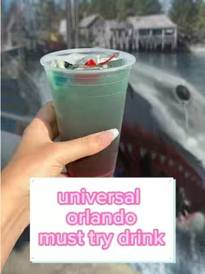 A hand holding a colorful cocktail in a clear plastic cup with a lid. The drink appears to be layered with red at the bottom, a green-blue in the middle, and topped with white. Behind the drink, a blurred background shows what appears to be a shark's mouth with teeth. Below the drink is a white rectangle with text in pink, stating 'universal orlando must try drink'.