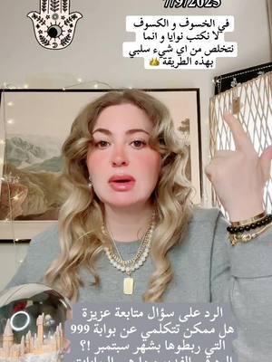 A woman with blonde hair is centered in the frame, looking directly at the camera and gesturing with her right hand. The background includes framed artwork and decorative elements. Text overlays in Arabic are present on the top of the image and at the bottom of the image. The overall composition suggests a video message or presentation.