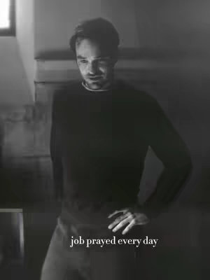 A black and white image featuring a man in a dark long-sleeved shirt, with his right hand on his hip. Soft lighting suggests an indoor setting.