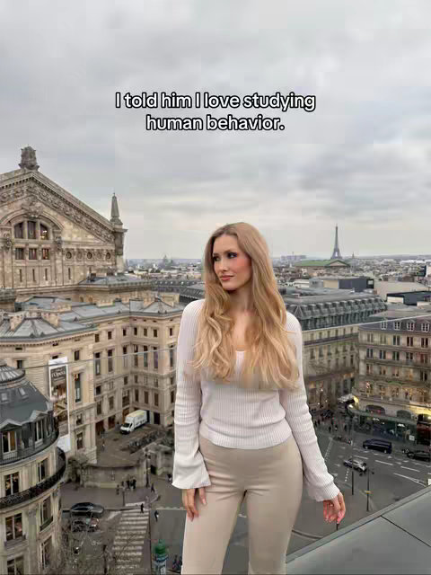 A woman stands on a rooftop overlooking a cityscape, with a text overlay above her. The background features buildings, a cathedral, and the Eiffel Tower in the distance. The woman is fair-skinned, with long, blonde hair, wearing a white sweater and beige pants. The overall tone is bright and the sky is overcast.