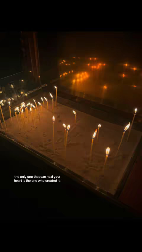 The image presents a dimly lit interior, likely a chapel or church, with an array of lit candles. The candles are positioned in a sandy tray, with a dark background that contrasts the candle flames and creates an impression of depth. Text overlayed near the bottom displays a spiritual message.