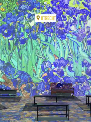 The image showcases the interior of the Van Gogh: The Immersive Experience, featuring a projection of Irises on a large wall. The immersive art installation is filled with vivid blues, greens, and yellows. In front of the projection are black benches and tables, giving the appearance of a waiting area or viewing space. A white location tag in the upper-left corner of the image, reading "UTRECHT". 