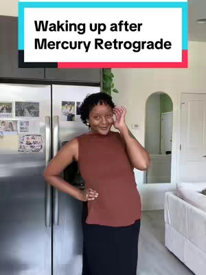 A woman with dark short curly hair stands in a modern kitchen, wearing a rust-colored top and a black skirt. The text "Waking up after Mercury Retrograde" is displayed in a white box with teal and red accents above her. She has a hand near her ear and appears to be listening or reacting to something. The background includes a stainless steel refrigerator, a light-colored wall, and glimpses of a living area with a white couch.