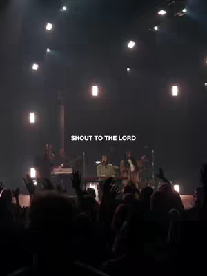 A dimly lit image of a worship service. A stage is in the middle, featuring people singing and playing instruments. The crowd in front has their hands raised. Large rectangular lights are visible in the dark background. The text 'Shout to the Lord' is overlaid near the top.