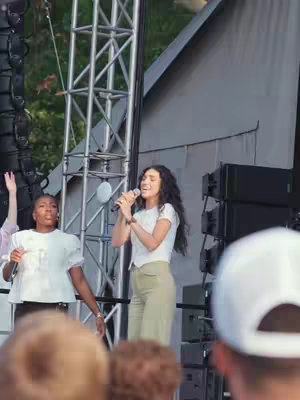 The cover image is a live shot from a concert or event. A woman with long, wavy dark hair wearing a white shirt and light green pants is singing into a handheld microphone. A second person holding a microphone is to her left, close-up. In the background are stage elements, structures, and other people, with an overcast sky.